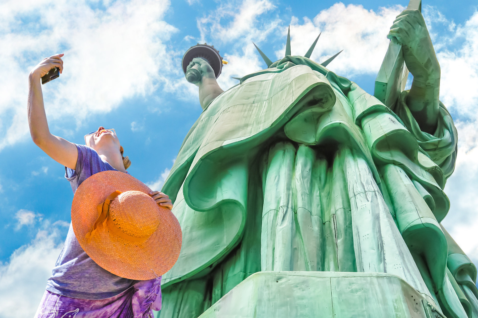 A smiling and fashionable woman with a orange wide-brimmed hat takes a selfie. Statue of Liberty and blue sky in the background. Liberty Island, New York City, United States.
