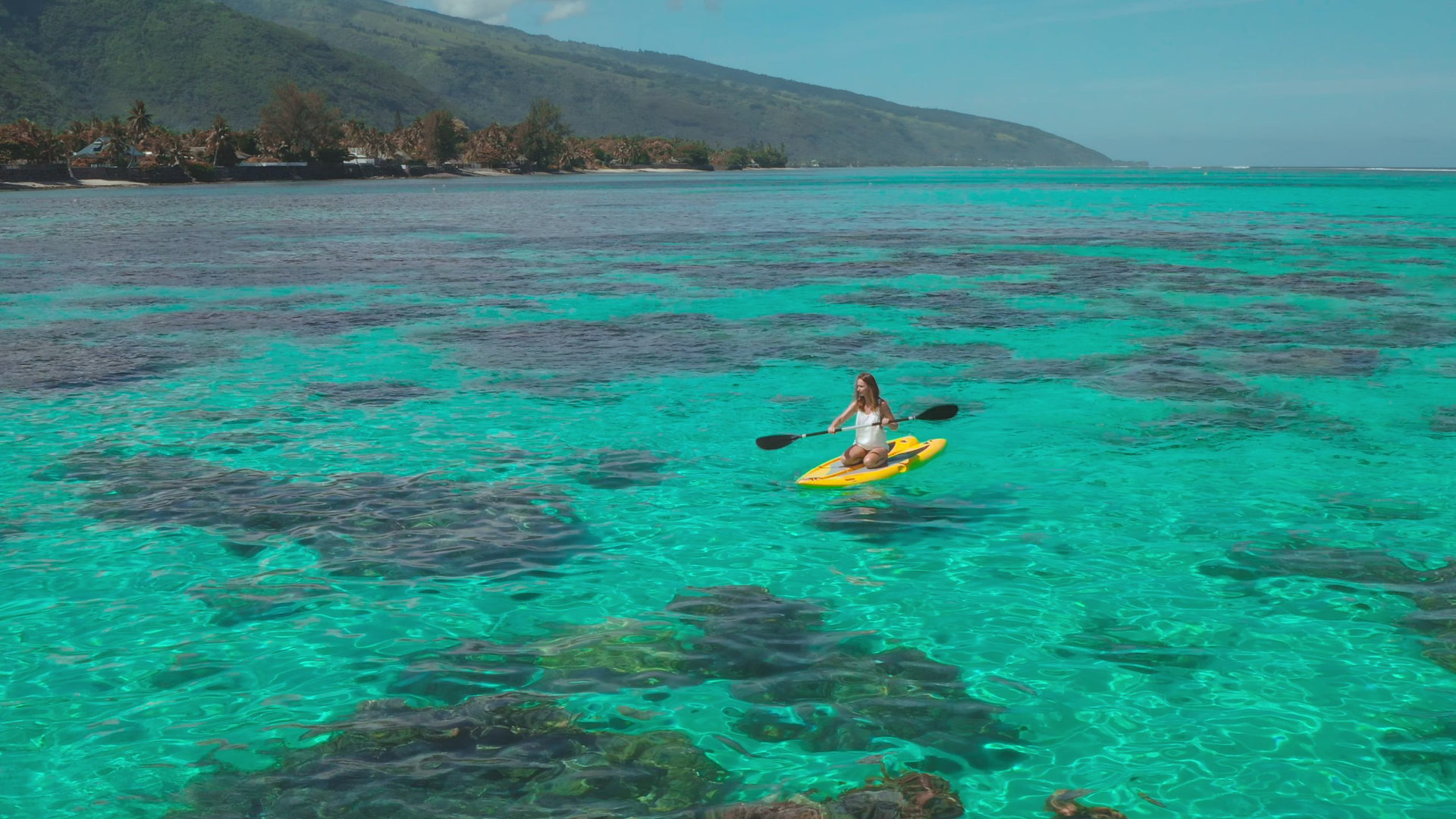 Aerial view of a woman kayaking over a coral reef in turquoise water near a tropical island. She is enjoying a sunny day in paradise