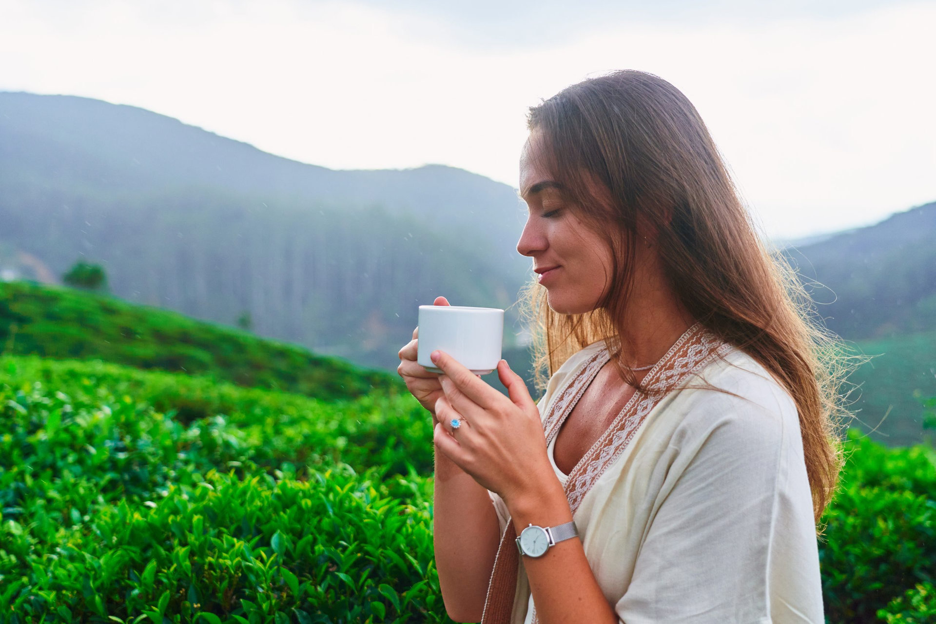 Woman traveler holding in hands and enjoying a cup of aromatic black tea against the background of a tea plantation while traveling in beautiful destination in Nuwara Eliya in Sri Lanka, Ceylon Island