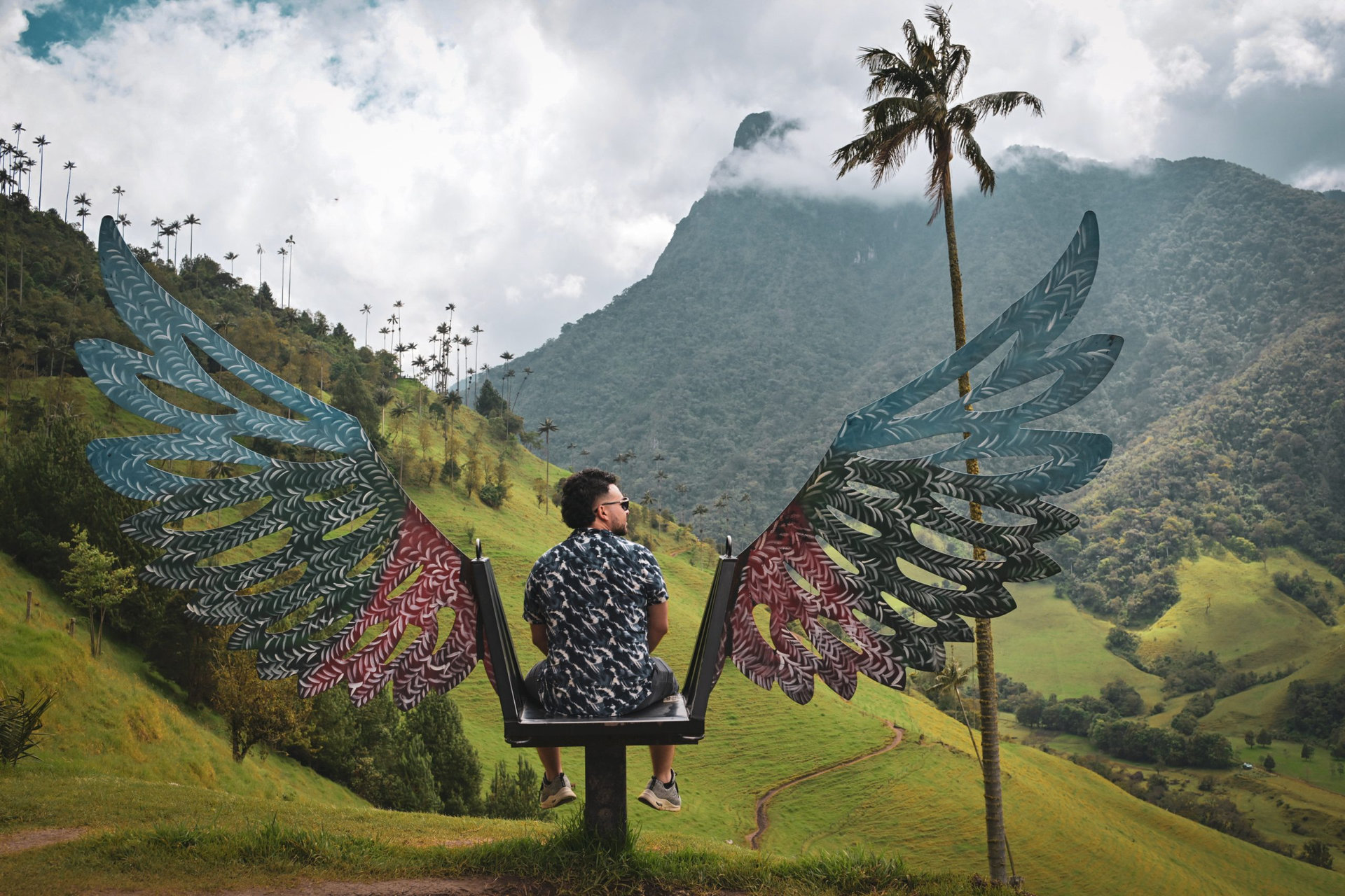 A man sits on a colorful winged chair overlooking Colombia's Cocora Valley. Surrounded by wax palms and mountains, it highlights adventure, nature, and cultural exploration in South America