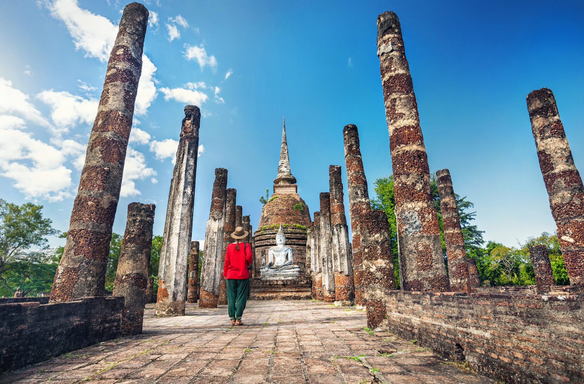 Tourist Woman in red costume looking at ancient Buddha statue in Wat Sa Si of Sukhothai Historical Park, Thailand