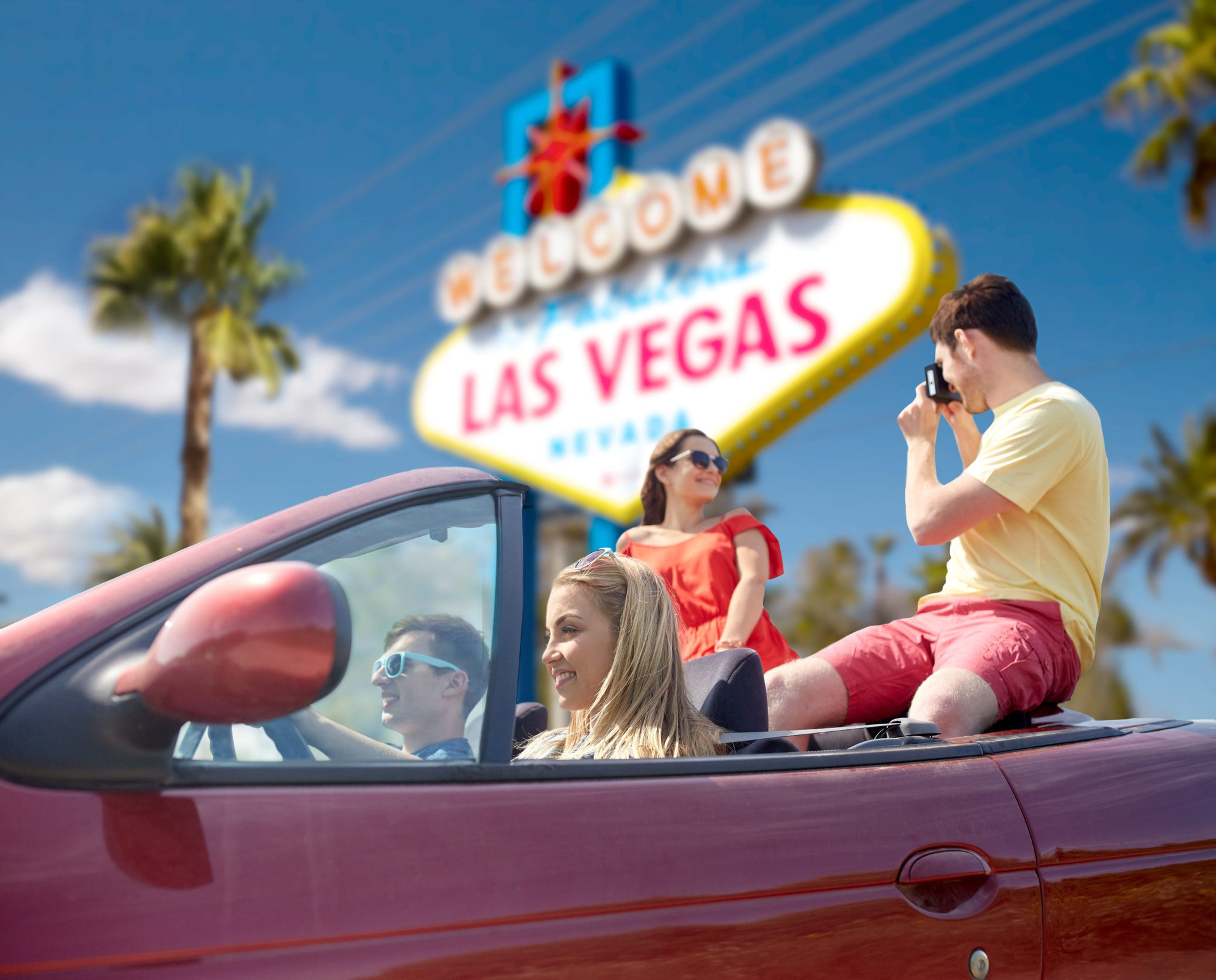 summer holidays, road trip and travel concept - happy friends driving in convertible car and taking picture by film camera over welcome to fabulous las vegas sign background
