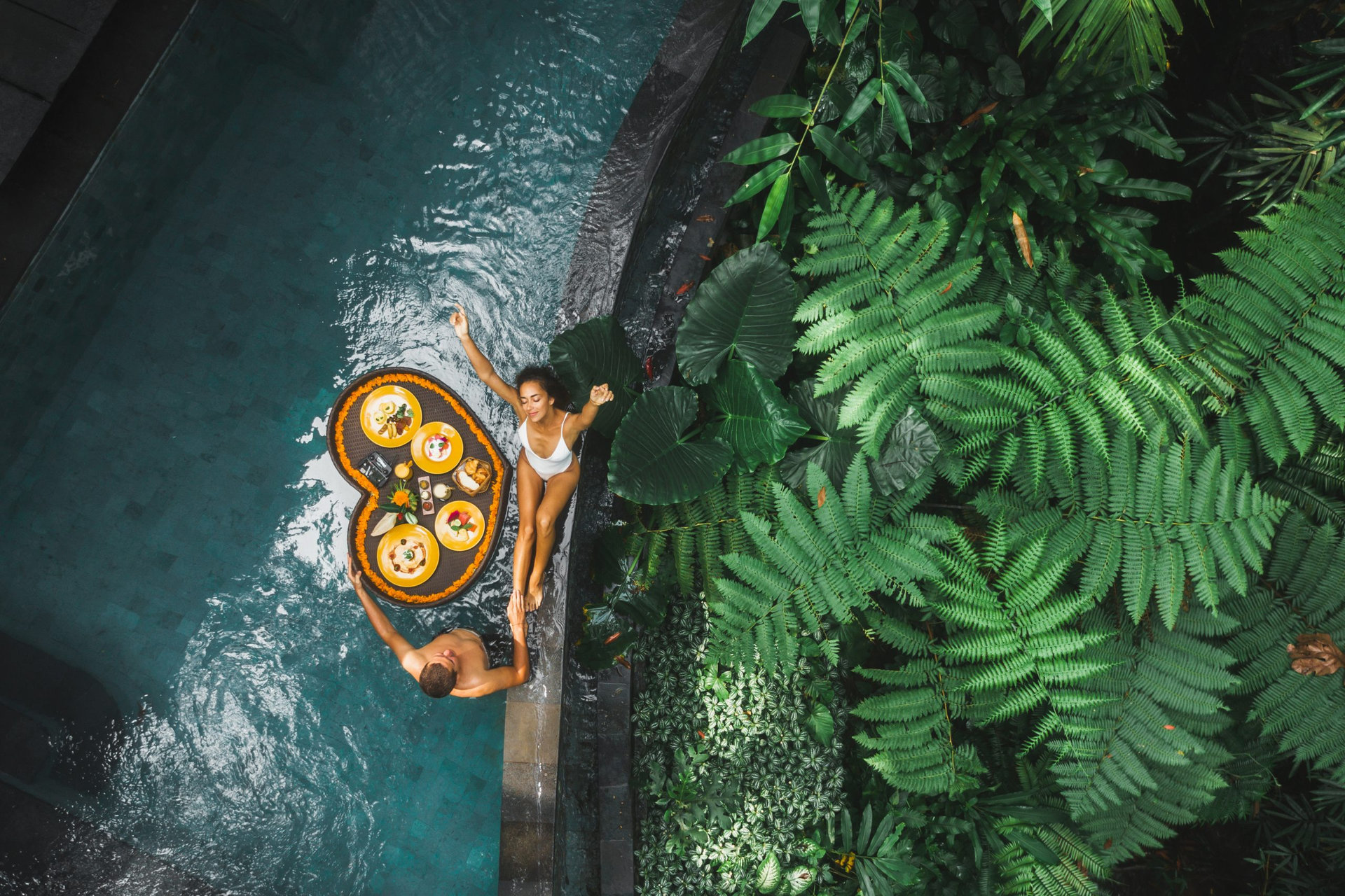 Travel happy couple in love eating floating breakfast in jungle swimming pool. Awakening in morning. Black rattan tray in heart shape, Valentines day or honeymoon surprise, view from above.