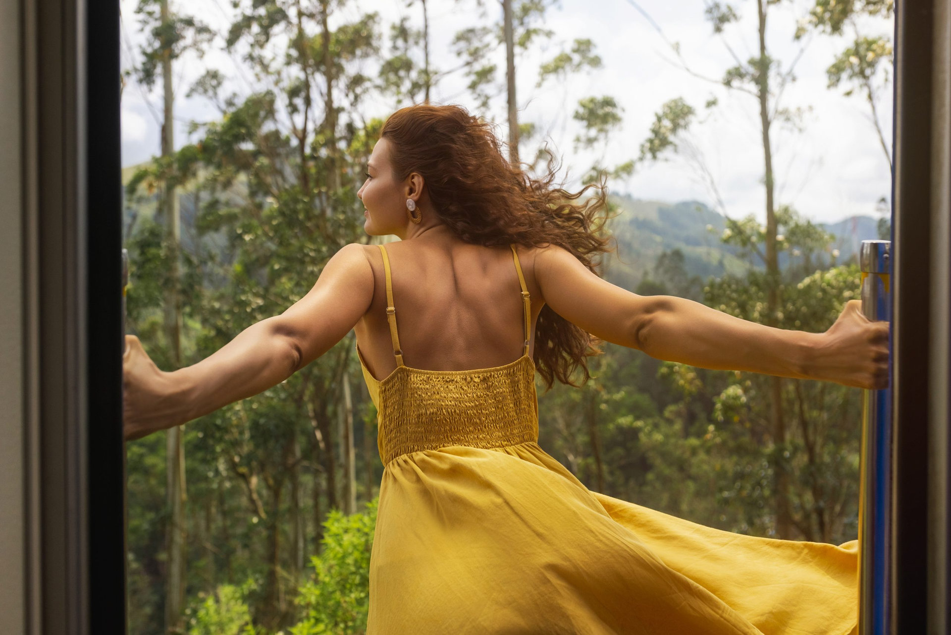 Travel and exploration of tourist woman by train to famous landmarks in Sri Lanka. Touristic journey around tropical country. Tour from Nuwara Eliya to Ella. Extreme attraction, brave passenger girl in dress standing out of the entrance of wagon with open doors.