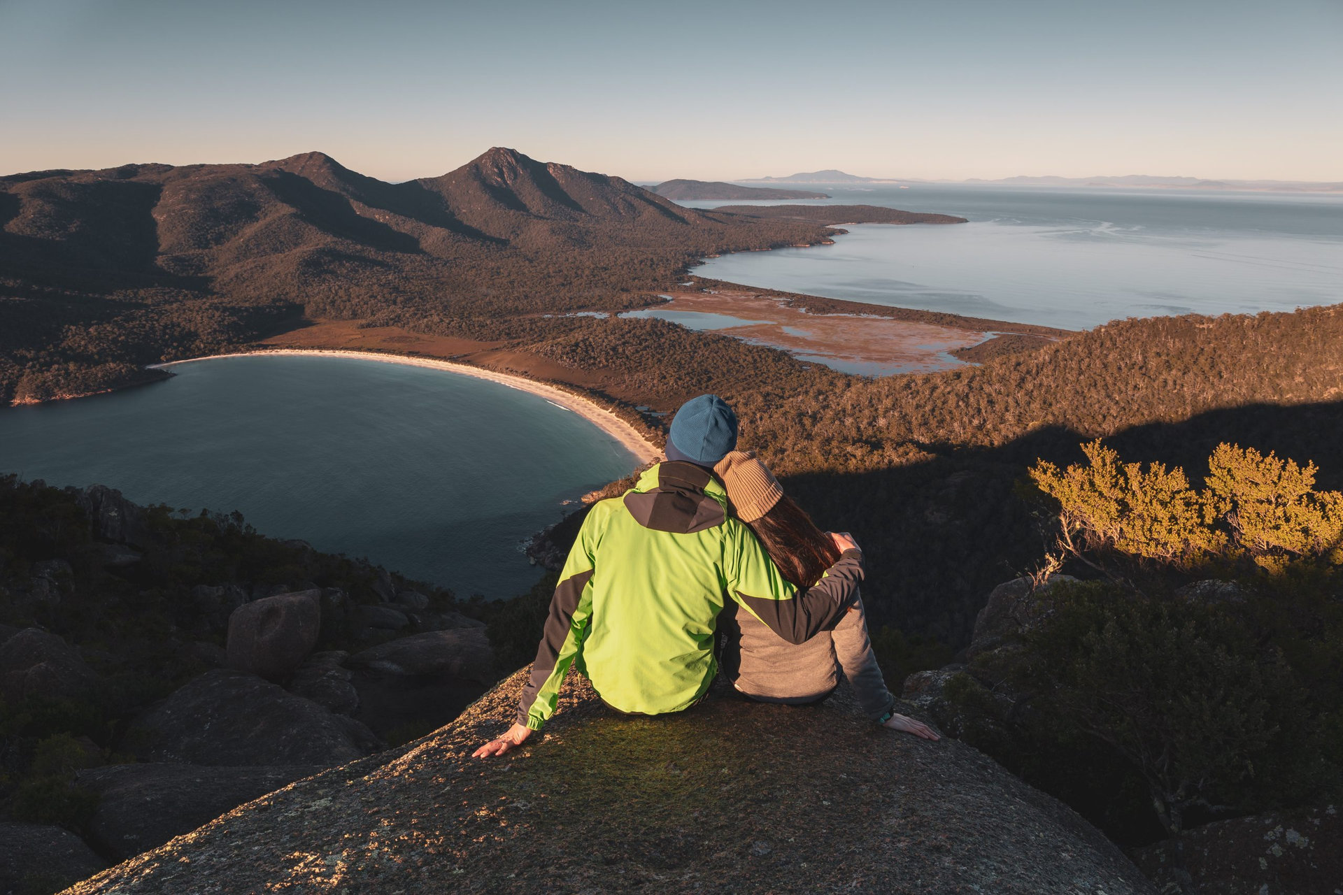 A pair of traveller couple watching sunrise at the peak of Mount Amos with Wineglass Bay at the background, Freycinet National Park, Coles Bay, Tasmania, Australia