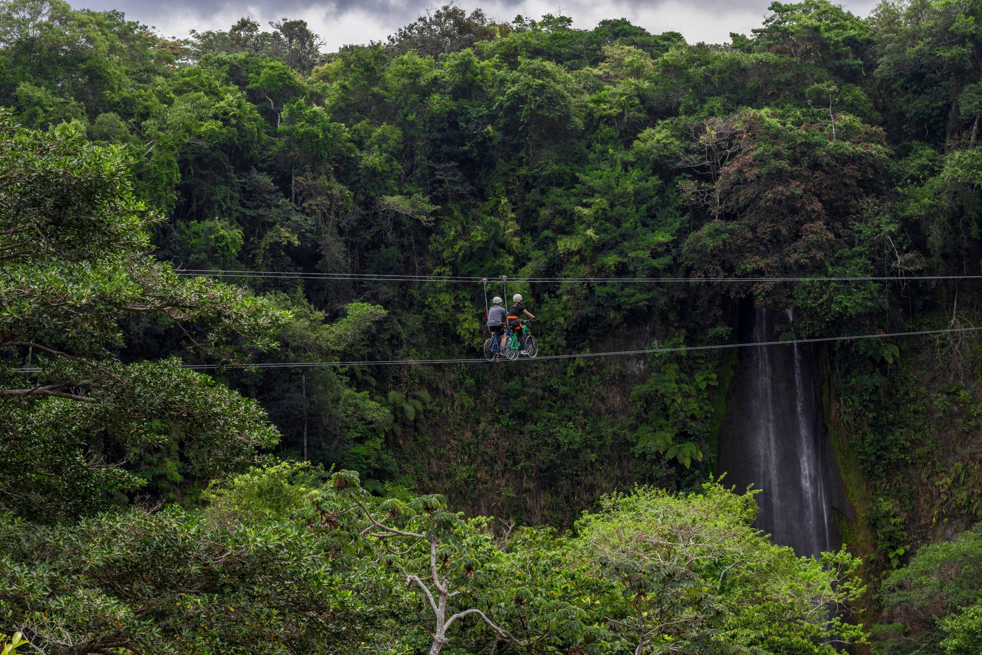 couple of hikers with harness and helmet riding zipline bicycles surrounded by vegetation and a beautiful waterfall in Costa Rica